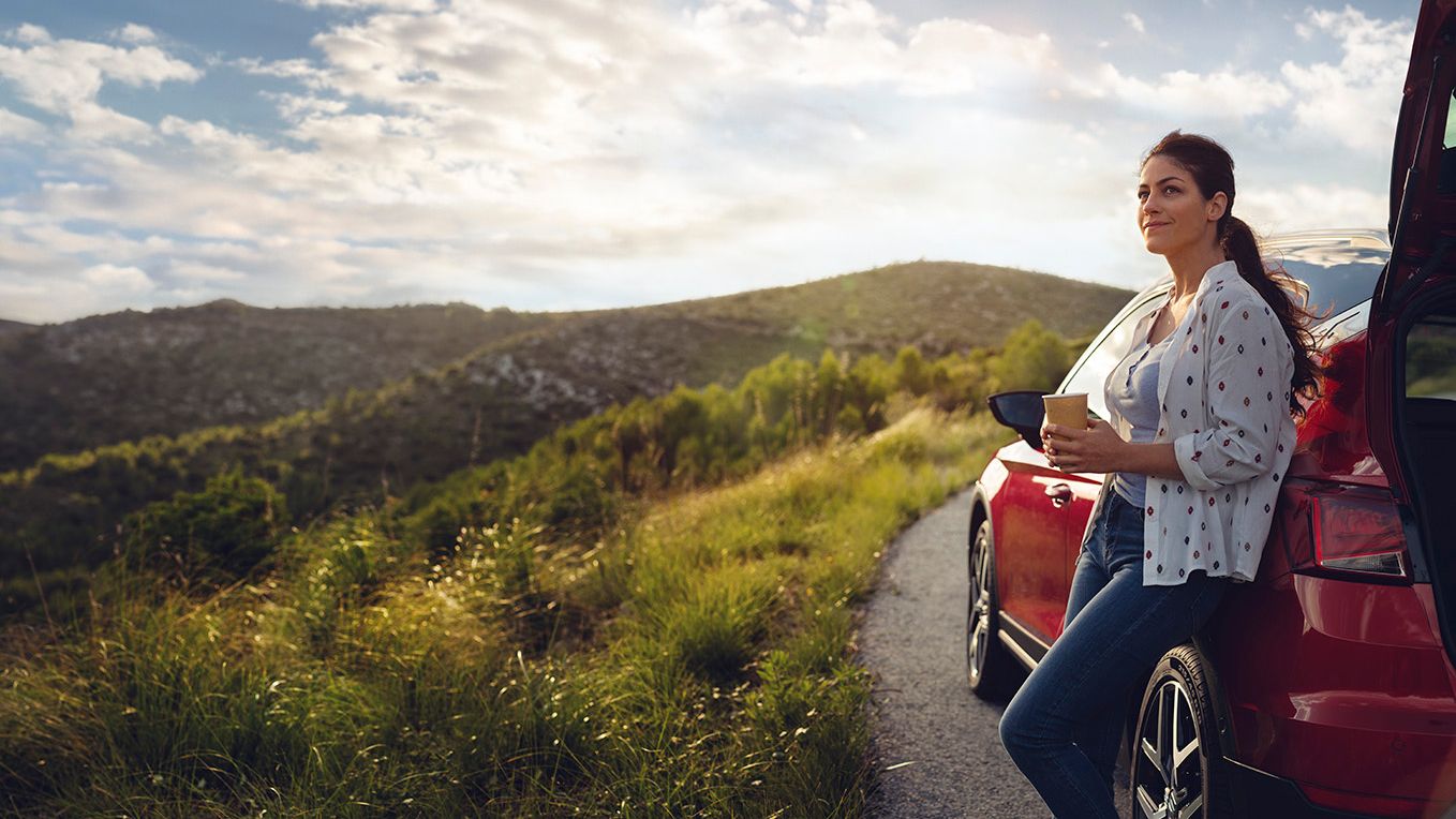 Eine Frau lehnt an einem roten SEAT Ibiza, hält einen Becher in der Hand und blickt auf die umliegende Berglandschaft entlang einer Landstraße.
